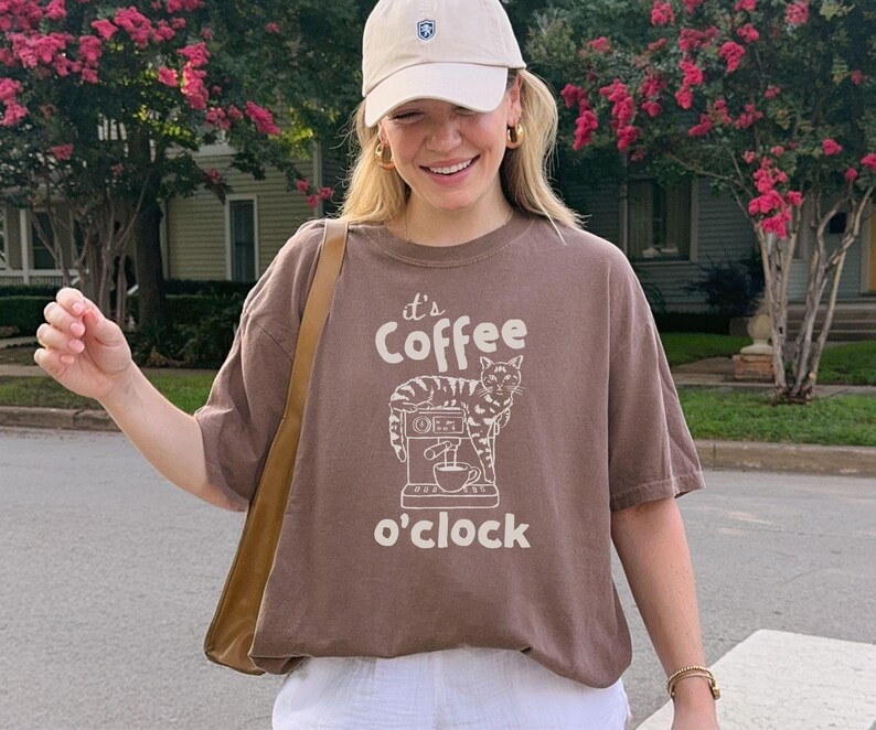 May include: Brown t-shirt with the text "It's Coffee o'clock" and a graphic of a cat and coffee machine. The shirt is worn with a beige cap, gold hoop earrings, and a brown shoulder bag. The person is smiling.