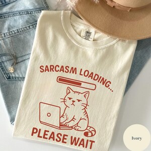 May include: Ivory-colored t-shirt with the text "Sarcasm Loading... Please Wait" in red. A cartoon cat with a grumpy expression is depicted in red, sitting in front of a laptop. The shirt is made of a soft material.