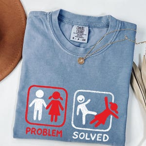 Puede incluir: Camiseta Comfort Colors azul claro con un gráfico que dice "Problema resuelto". El gráfico muestra una pareja de figuras de palo en un cuadro rojo y una figura de palo pateando una figura roja en un cuadro blanco. Un collar dorado está sobre la camisa.