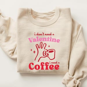 May include: A cream-coloured sweatshirt with the text "i don't need a Valentine i need a Coffee" in pink and red. The graphic shows a hand holding a coffee cup. The sweatshirt is folded.