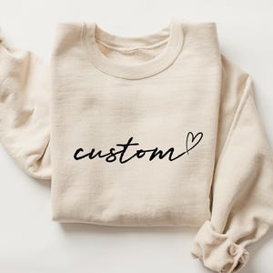 May include: A cream-colored sweatshirt with the word "custom" in black script, followed by a heart symbol. The sweatshirt is folded, with the sleeves visible on either side.