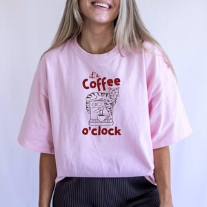 May include: Light pink t-shirt with the text "It's Coffee o'clock" in red. The graphic features a cat standing next to a coffee machine. The shirt has short sleeves and a relaxed fit.