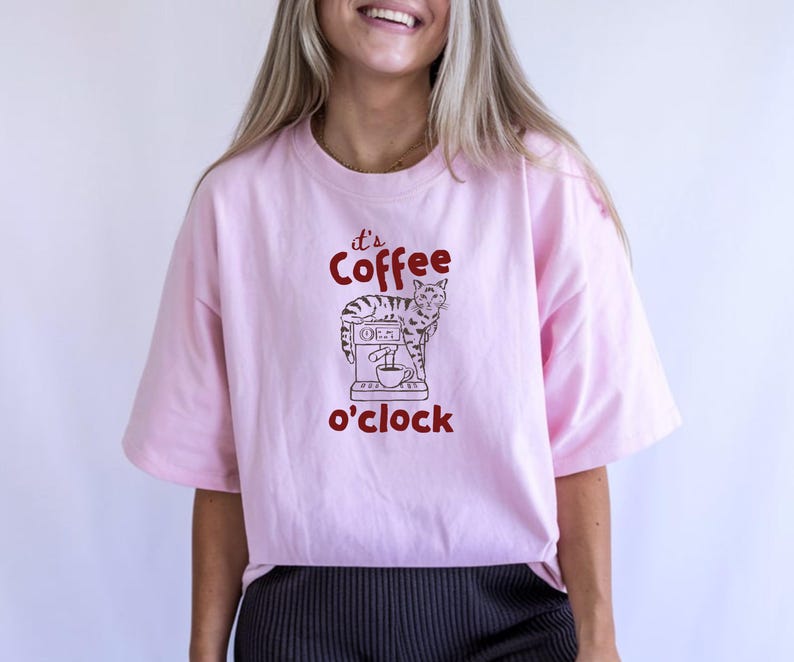 May include: Light pink t-shirt with the text "It's Coffee o'clock" in red. The graphic features a cat standing next to a coffee machine. The shirt has short sleeves and a relaxed fit.