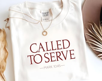 Può includere: T-shirt color crema con la scritta "CALLED TO SERVE" in bordeaux, con "MARK 10:45" sotto. Una collana d'oro con un ciondolo circolare è appoggiata sulla maglietta. La maglietta ha un'etichetta con la scritta "COUNTRY COLORS".