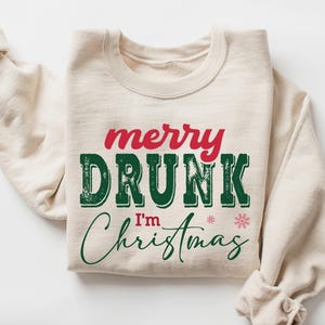 Könnte beinhalten: Beiges Sweatshirt mit dem Text "merry DRUNK I'm Christmas" in Rot und Grün. Neben dem Wort "Christmas" befindet sich ein kleiner rosa Schneeflocke. Das Sweatshirt hat lange Ärmel und einen Rundhalsausschnitt.