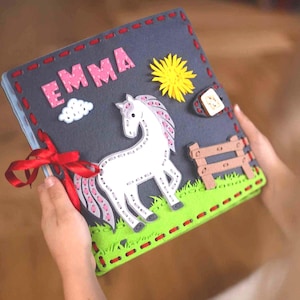 Op de afbeelding: Een vierkant vilten activiteitenboek met een donkergrijze kaft met de naam EMMA in roze letters. De kaft toont een wit paard met roze manen, een gele zon, een witte wolk en een bruin hekwerk op een basis van groen gras. Een rood lint is aan de zijkant.