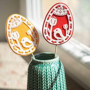 Può includere: Due decorazioni a forma di uovo di Pasqua su bastoncini di legno, una gialla e una rossa, ciascuna con un uccello bianco e un motivo floreale. Le uova sono in un vaso turchese con un motivo strutturato, perfette per l'arredamento primaverile.