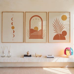 Set of 3 boho wall art prints featuring terracotta moon phases, abstract sun arch, and botanical palm leaf illustrations framed symmetrically on a warm sunlit wall above a floating wooden shelf with decorative items in a modern living room.