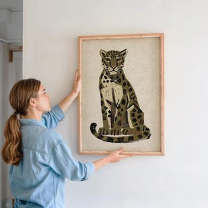 A woman in a denim shirt holds a framed print of a sitting leopard with dark green and brown spots on a light background, against a light wall.