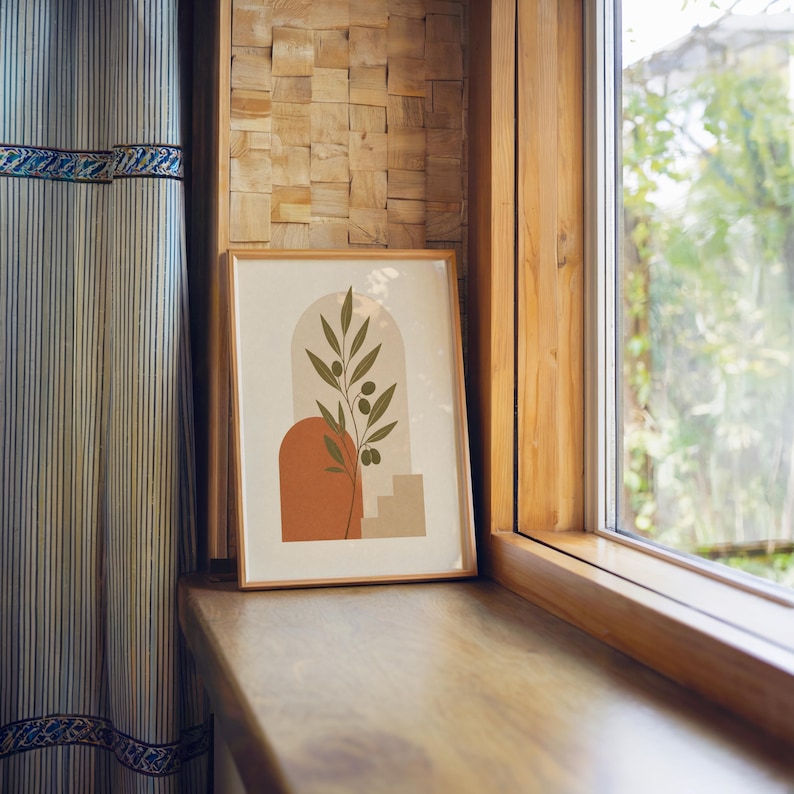 Single framed olive branch art print resting near a window with warm sunlight and curtains.