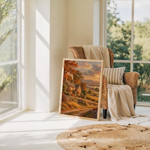 Autumn cottage landscape wall art print leaning on the floor beside a rattan armchair and jute rug in a bright sunlit room.