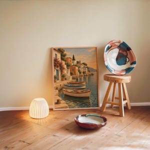Mediterranean coastal village wall art print leaning against a warm beige wall on a wooden floor beside a stool and a glowing lamp.