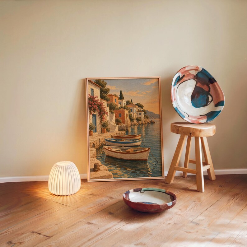 Mediterranean coastal village wall art print leaning against a warm beige wall on a wooden floor beside a stool and a glowing lamp.