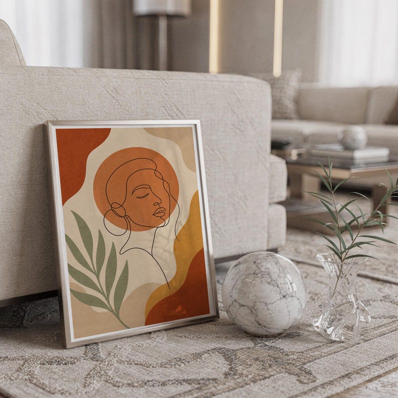 Abstract line art face print in a silver frame leaning against a sofa beside a decorative marble sphere.