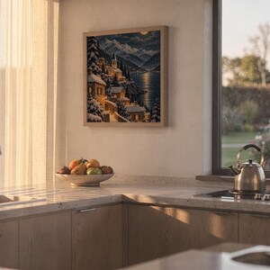 Snowy mountain village wall art print hanging above a stone countertop with a fruit bowl in a sunny kitchen.