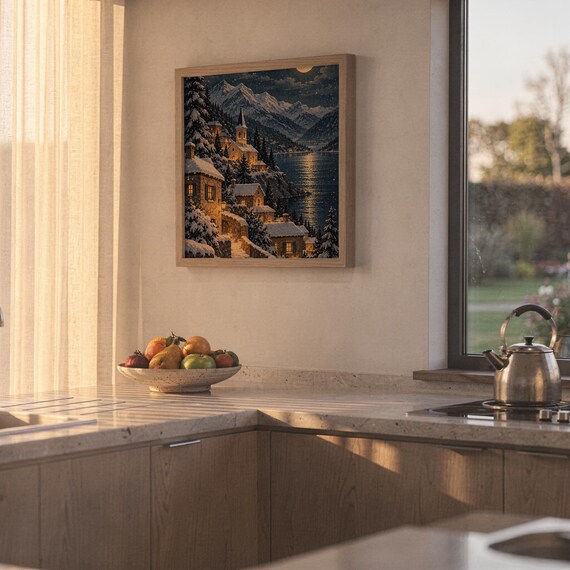 Snowy mountain village wall art print hanging above a stone countertop with a fruit bowl in a sunny kitchen.