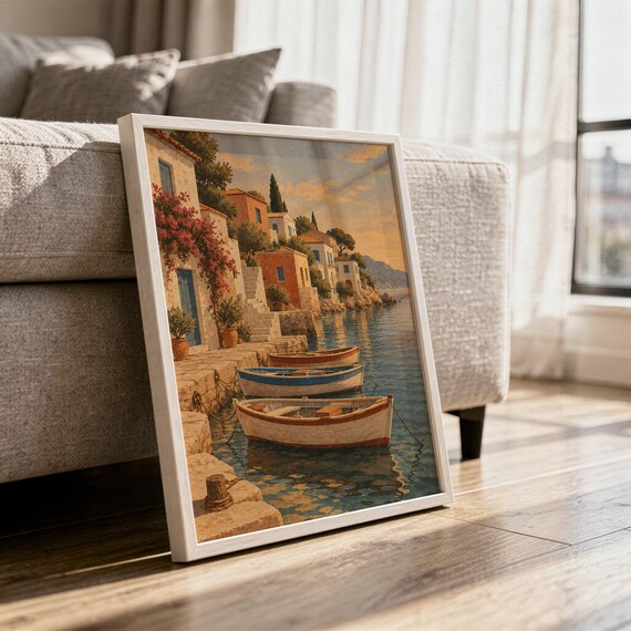 Mediterranean coastal village wall art print in a white frame leaning against a gray sofa on a sunlit wooden floor.