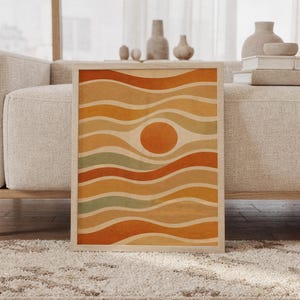Abstract wavy sun wall art print of a wavy bands around a central sun circle in burnt orange, mustard, tan, cream and sage green, framed and leaning against a beige sofa on a textured rug in a bright Scandinavian living room.