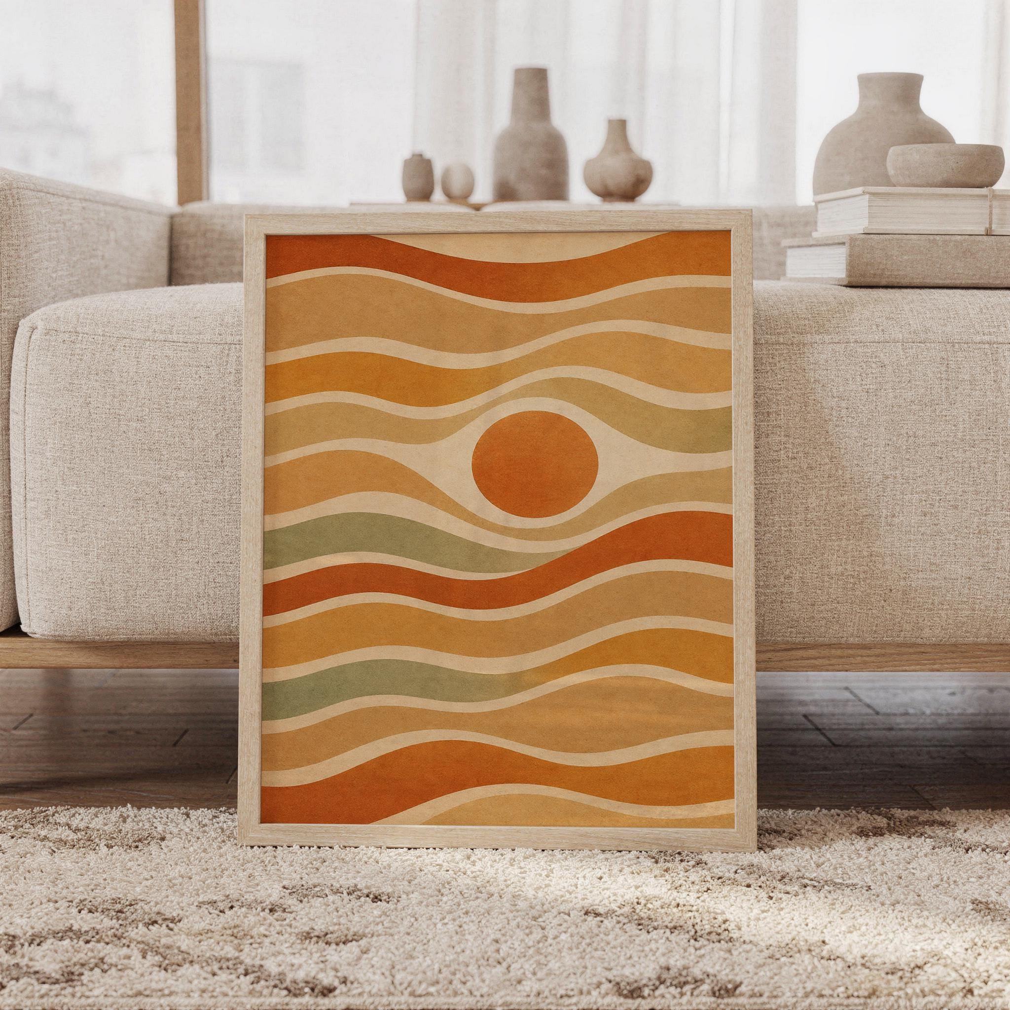 Abstract wavy sun wall art print of a wavy bands around a central sun circle in burnt orange, mustard, tan, cream and sage green, framed and leaning against a beige sofa on a textured rug in a bright Scandinavian living room.