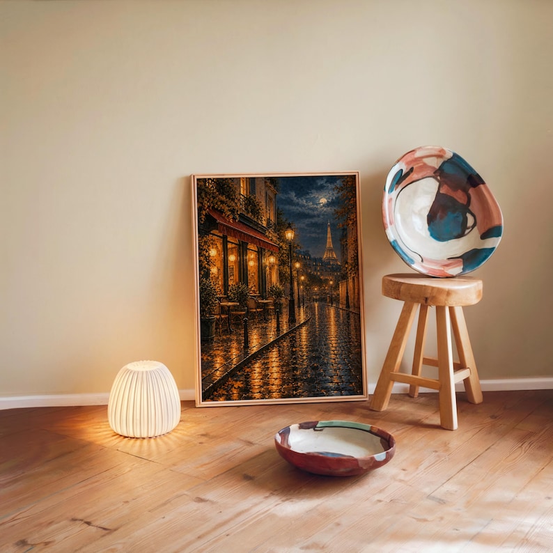 Paris eiffel tower wall art print leaning against a warm beige wall on a wooden floor beside a stool and a glowing lamp.