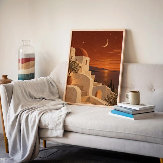 Mediterranean moonlit coastal wall art print leaning on a cream sofa against a beige wall beside books and a mug.