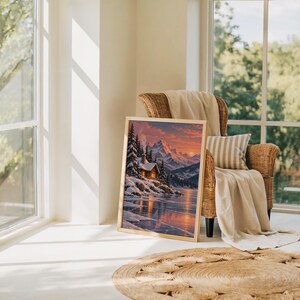 Winter cabin mountain wall art print leaning on the floor beside a rattan armchair and jute rug in a bright sunlit room.
