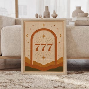 May include: A framed rectangular wall art print featuring the number 777 in a bold, dark orange font centered within a large archway. The design includes radiating sunburst lines, small stars, and crescent moons against a light beige background.
