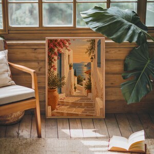 Mediterranean coastal village wall art print leaning against warm wood paneling beside a wooden armchair and a monstera plant.