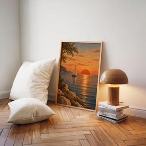 Sunset sailboat wall art print leaning on a herringbone wood floor beside cream cushions and a wooden mushroom lamp.