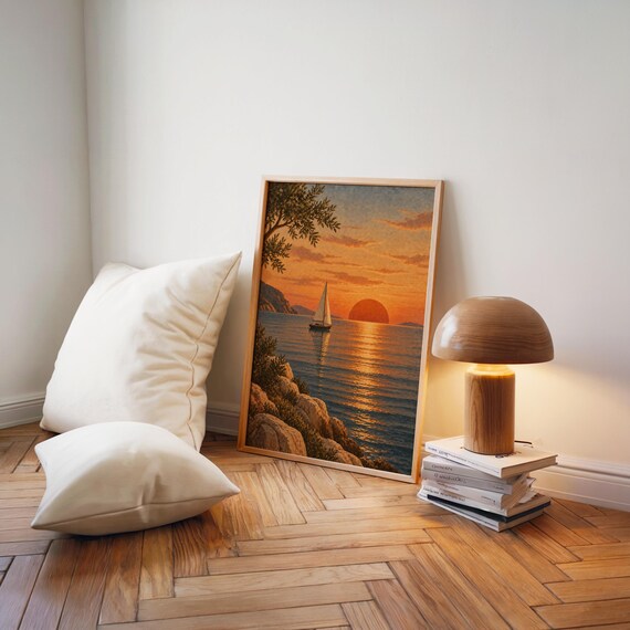 Sunset sailboat wall art print leaning on a herringbone wood floor beside cream cushions and a wooden mushroom lamp.
