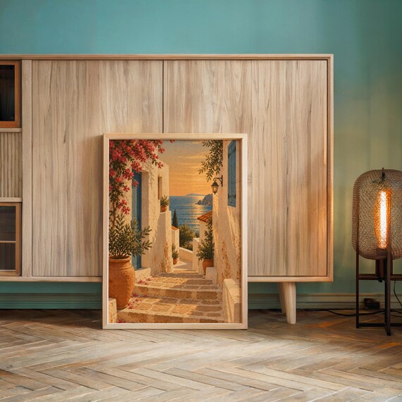 Mediterranean coastal village wall art print leaning against a wooden cabinet by a teal wall beside a glowing floor lamp.