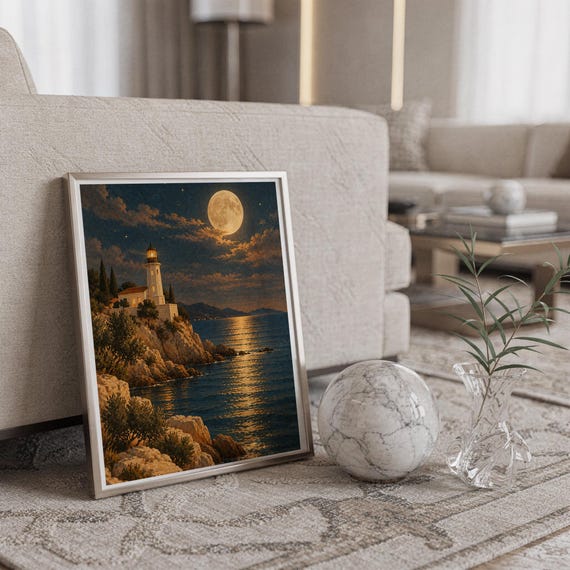 Moonlit lighthouse wall art print in a silver frame leaning against a sofa beside a decorative marble sphere.