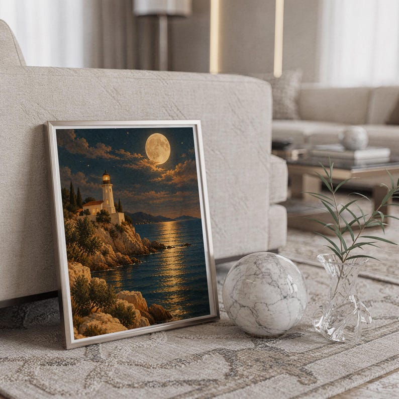 Moonlit lighthouse wall art print in a silver frame leaning against a sofa beside a decorative marble sphere.