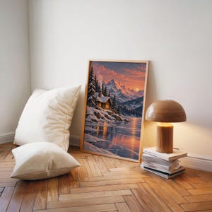Winter cabin mountain wall art print leaning on a herringbone wood floor beside cream cushions and a wooden mushroom lamp.