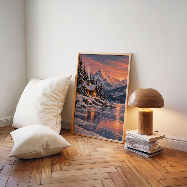 Winter cabin mountain wall art print leaning on a herringbone wood floor beside cream cushions and a wooden mushroom lamp.