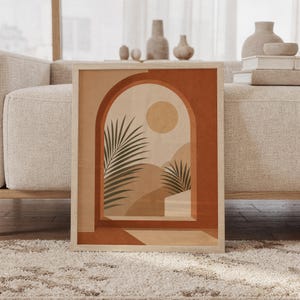 Framed terracotta arch window palm art print leaning on carpet in front of sofa in bright living room.