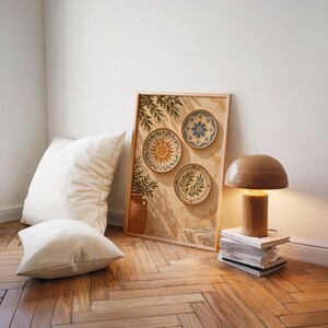 Mediterranean ceramic plate wall art print leaning on a herringbone wood floor beside cream cushions and a wooden mushroom lamp.
