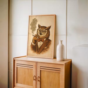 A framed print of an owl in a brown suit, red bow tie, and glasses, smoking a cigar with smoke curling up, sits on a light wood shelf with a pink salt lamp and a brown vase.