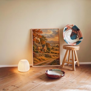 Autumn cottage landscape wall art print leaning against a warm beige wall on a wooden floor beside a stool and a glowing lamp.