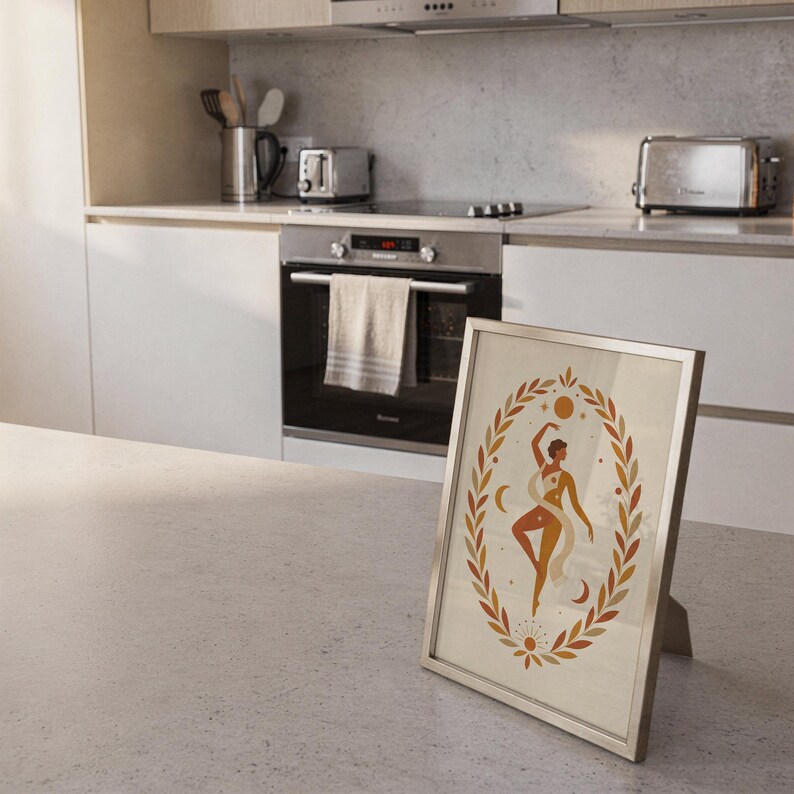 Framed terracotta divine feminine art print resting on the floor in a bright, modern kitchen interior.