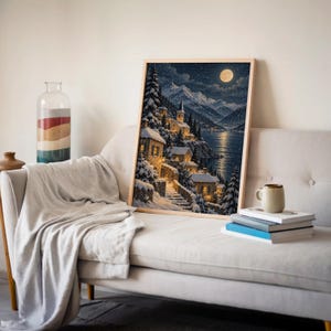 Snowy mountain village wall art print leaning on a cream sofa against a beige wall beside books and a mug.