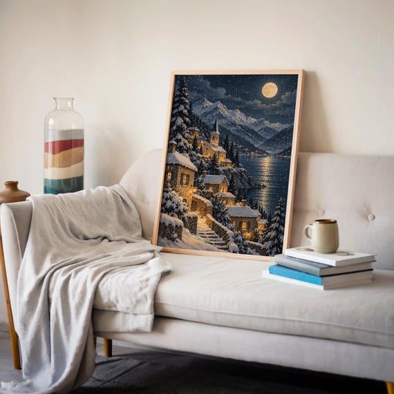 Snowy mountain village wall art print leaning on a cream sofa against a beige wall beside books and a mug.