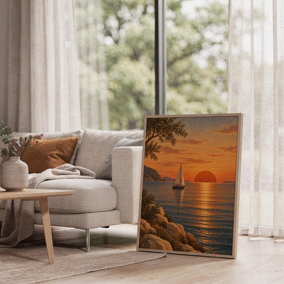 Sunset sailboat wall art print leaning on the floor beside a cream armchair next to large garden windows.