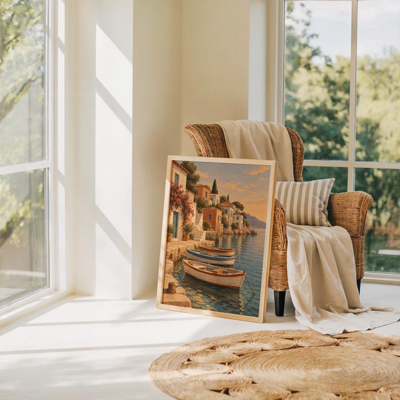 Mediterranean coastal village wall art print leaning on the floor beside a rattan armchair and jute rug in a bright sunlit room.