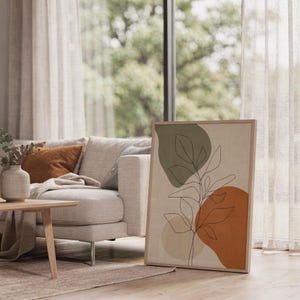 Framed sage green botanical artwork resting on the floor next to a grey sofa in a sunlit room.