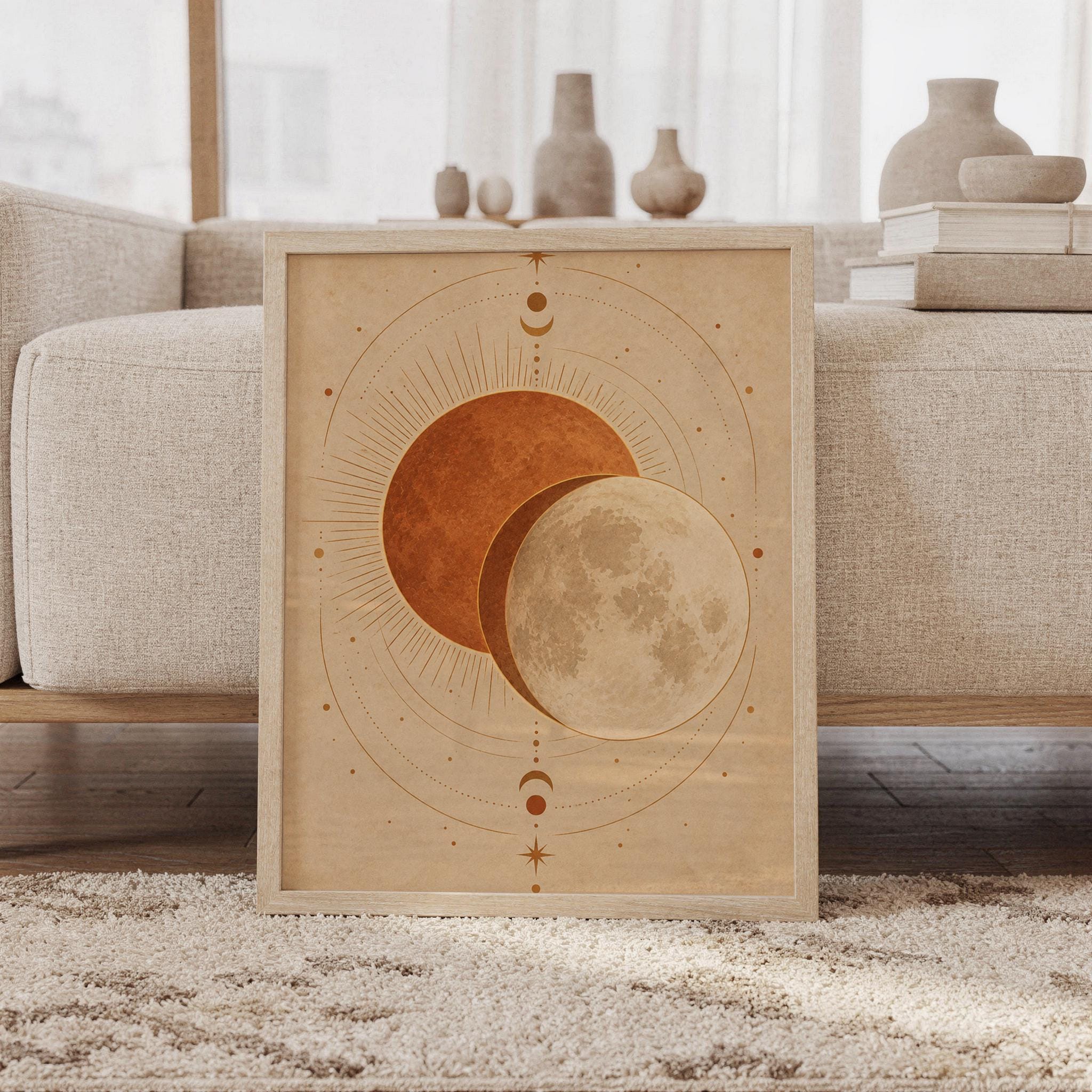 Sun and moon celestial wall art print featuring warm terracotta, ochre, cream, and parchment beige tones, layered sun and full moon imagery, celestial phases, stars, and delicate orbital lines, framed and leaning against a beige sofa on a textured rug in a bright Scandinavian living room.