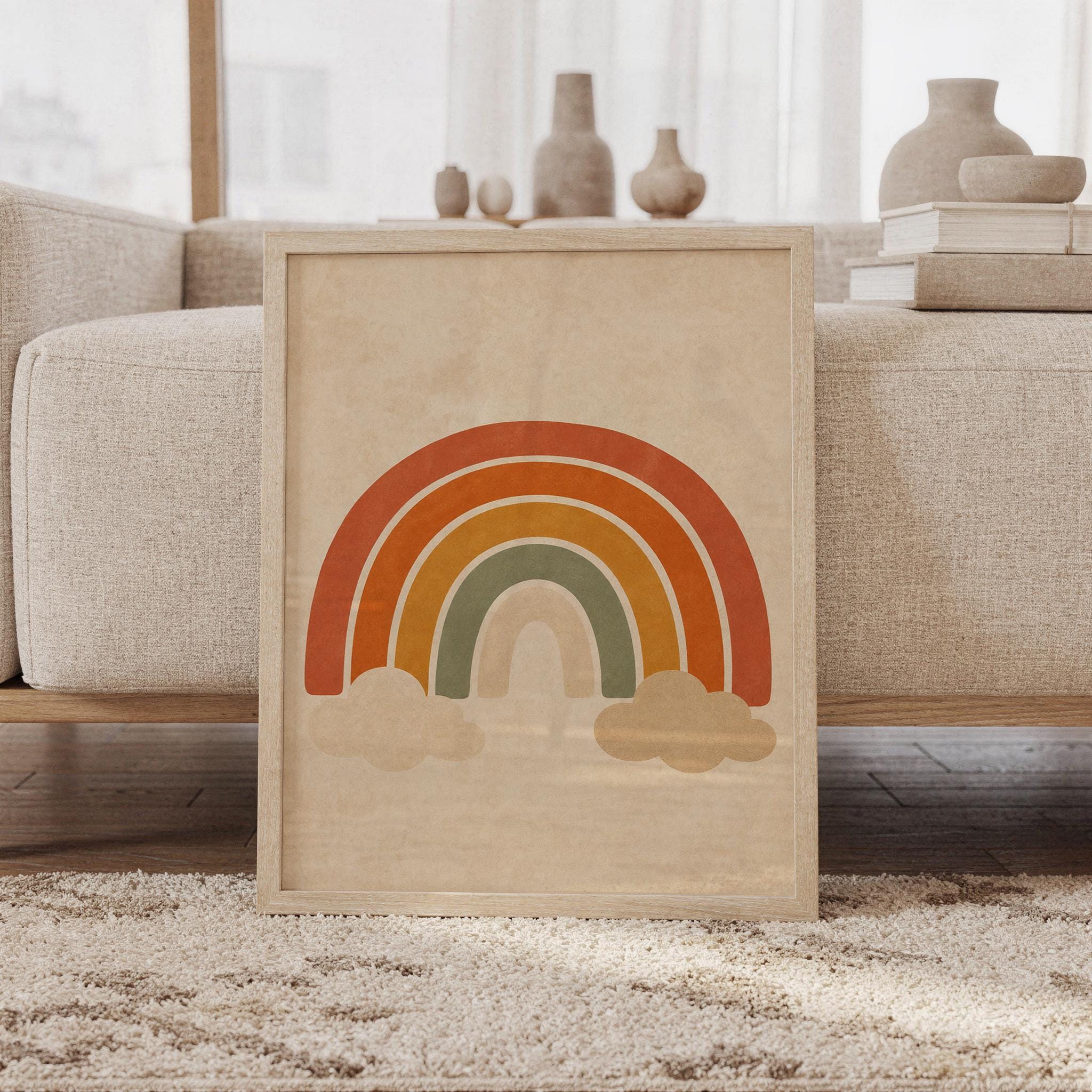 Boho rainbow wall art print of a layered rainbow arches with soft beige clouds in terracotta, burnt orange, mustard, sage and cream, framed and leaning against a beige sofa on a textured rug in a bright Scandinavian living room.