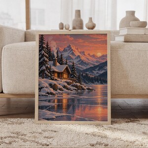 Winter cabin mountain wall art print featuring warm coral, peach, and slate blue sunset tones, a glowing log cabin, snow-covered pines, dramatic alpine peaks, and a frozen lake, framed and leaning against a beige sofa on a textured rug in a bright Scandinavian living room.