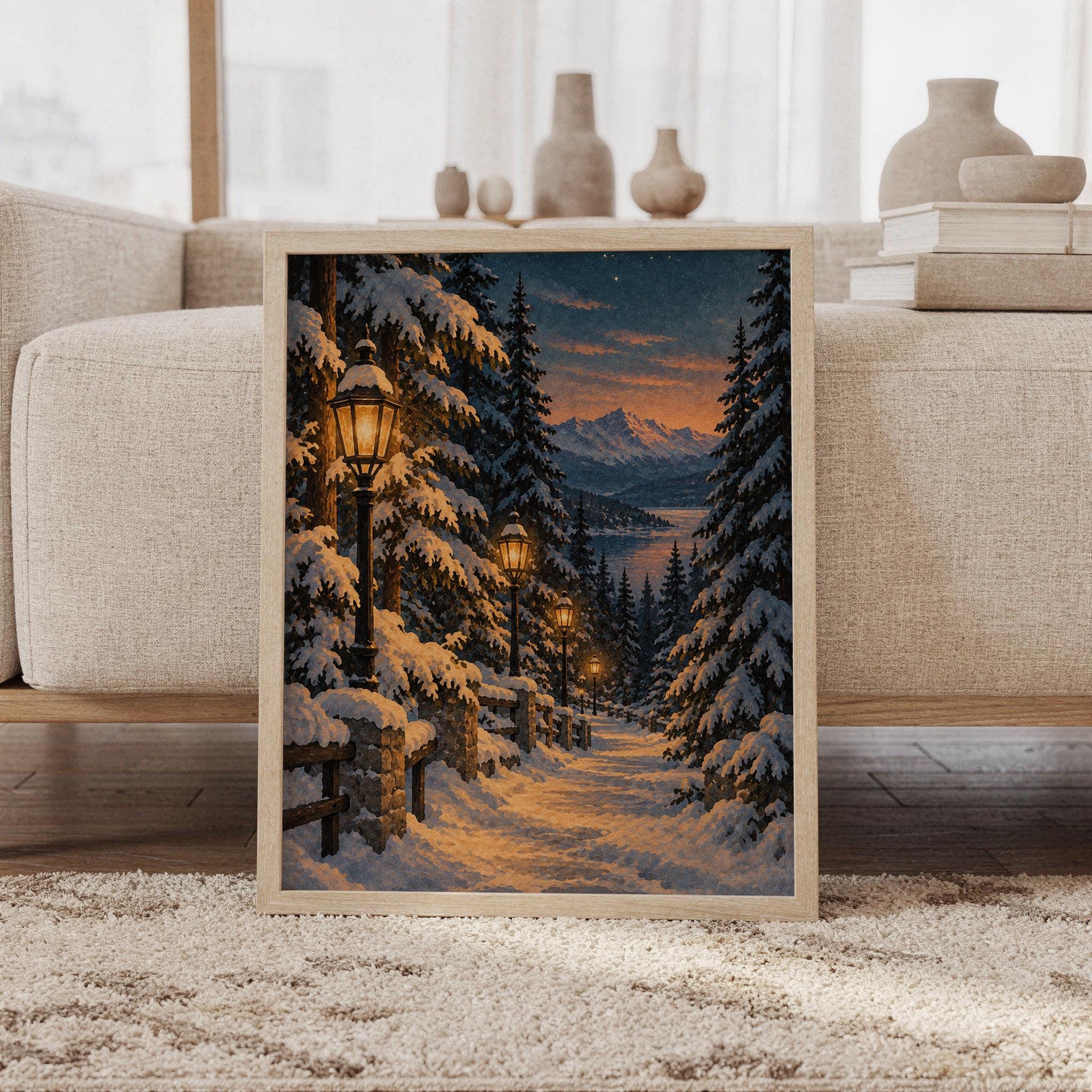 Snowy forest path wall art print featuring deep midnight blue skies, snow-covered pine trees, glowing golden lanterns, and distant mountain peaks in a painterly winter landscape style, framed and leaning against a beige sofa on a textured rug in a bright Scandinavian living room.