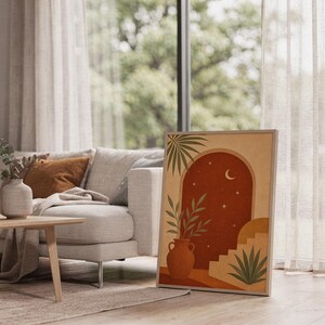 Terracotta moon and stars arch art print leaning on the floor beside a cream armchair next to large garden windows.